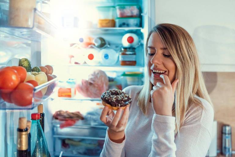 Mulher olhando para donut com a porta da geladeira aberta