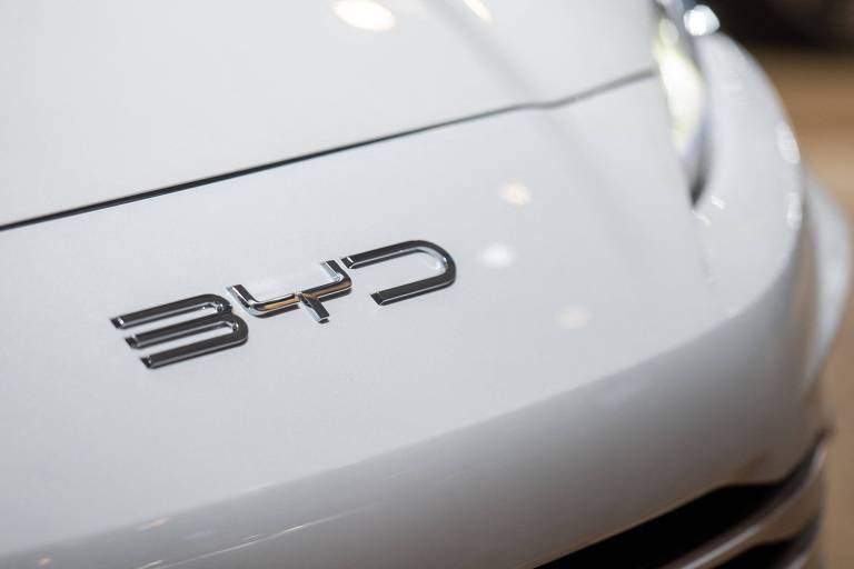 A imagem mostra a parte frontal de um carro branco， destacando o logotipo da marca BYD， que está localizado no capô. O logotipo é em letras prateadas e está posicionado à esquerda da imagem.