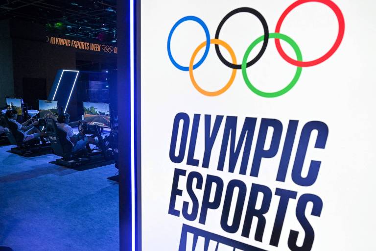 A imagem mostra um estande da Semana Olímpica de Esportes Eletrônicos， com um painel à direita exibindo o logotipo que inclui os anéis olímpicos coloridos e o texto 039;OLYMPIC ESPORTS WEEK039;. Ao fundo， há várias pessoas jogando em simuladores de corrida， com telas de jogos visíveis.