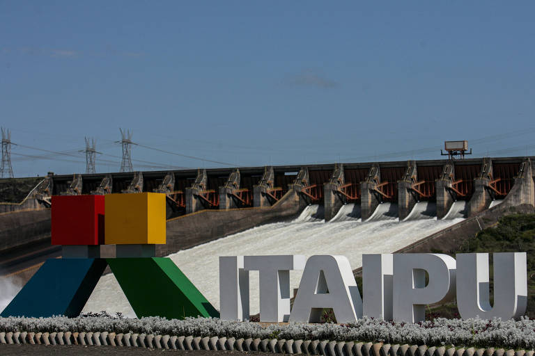 A imagem mostra a Usina Hidrelétrica de Itaipu, com a estrutura da barragem ao fundo e uma grande placa com a palavra 'ITAIPU' em letras brancas. Na parte superior da barragem, há várias comportas abertas, liberando água. No canto esquerdo da imagem, há um logotipo colorido composto por quadrados amarelo, vermelho e verde, e um triângulo azul.