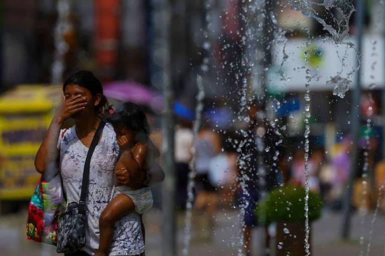 A imagem mostra uma mulher com uma criança nas costas， passando por uma fonte de água em uma área urbana. Ela está com a mão cobrindo o rosto， possivelmente para se proteger da água. Ao fundo， há várias pessoas e um veículo amarelo. A cena é ensolarada e há plantas em vasos ao redor.