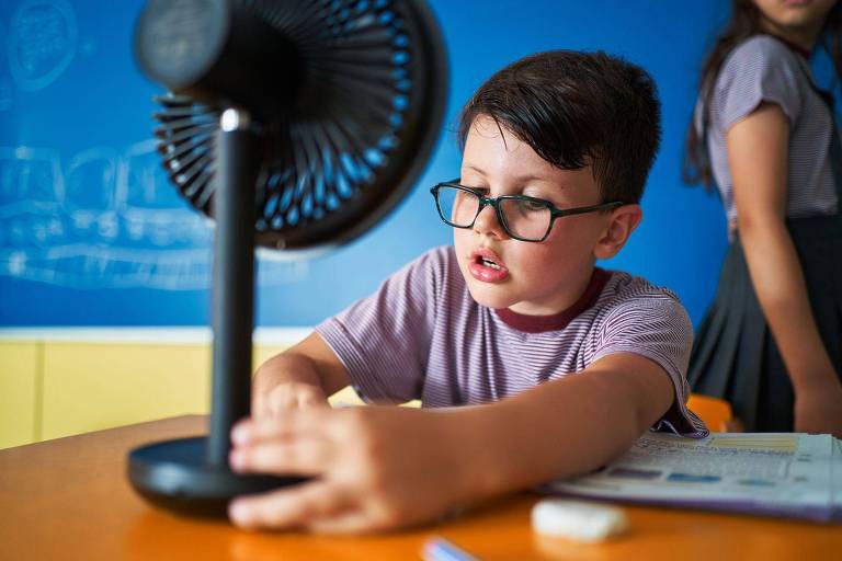 A imagem mostra um menino com óculos， concentrado em ajustar um ventilador de mesa. Ele está sentado em uma mesa， vestindo uma camiseta roxa. Ao fundo， há uma parede azul e uma menina em pé， parcialmente visível， vestindo uma saia. O ambiente parece ser uma sala de aula.