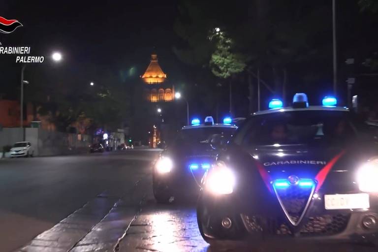 A imagem mostra dois carros da polícia， identificados como Carabinieri， estacionados em uma rua de Palermo à noite. As luzes azuis dos veículos estão acesas， iluminando a área ao redor. Ao fundo， é possível ver um edifício iluminado， que parece ser um monumento ou uma torre. A rua está deserta， com algumas árvores e postes de luz visíveis.