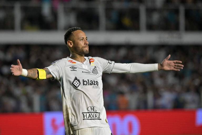 Um jogador de futebol， vestindo um uniforme branco com detalhes em preto e amarelo， está em campo com os braços abertos， em uma pose de celebração ou interação com a torcida. Ele usa uma faixa de capitão no braço e tem tatuagens visíveis. Ao fundo， há uma multidão de torcedores e um painel publicitário iluminado.