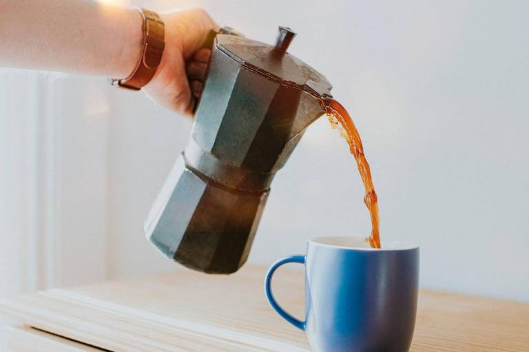 Uma mão está segurando uma cafeteira italiana preta， enquanto derrama café em uma caneca azul. O fundo é claro e a superfície onde a caneca está apoiada é de madeira.