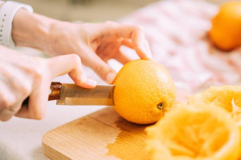 Uma mão segurando uma laranja enquanto a outra mão a corta com uma faca. Ao fundo， há mais laranjas e cascas de laranja em uma tábua de corte. O ambiente é claro e bem iluminado.