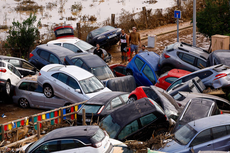 A imagem mostra uma cena de destruição causada por uma inundação， com vários carros empilhados e danificados em uma área alagada. Ao fundo， há algumas pessoas observando a situação. A água da inundação é visível ao redor dos veículos， e a vegetação nas proximidades parece afetada.