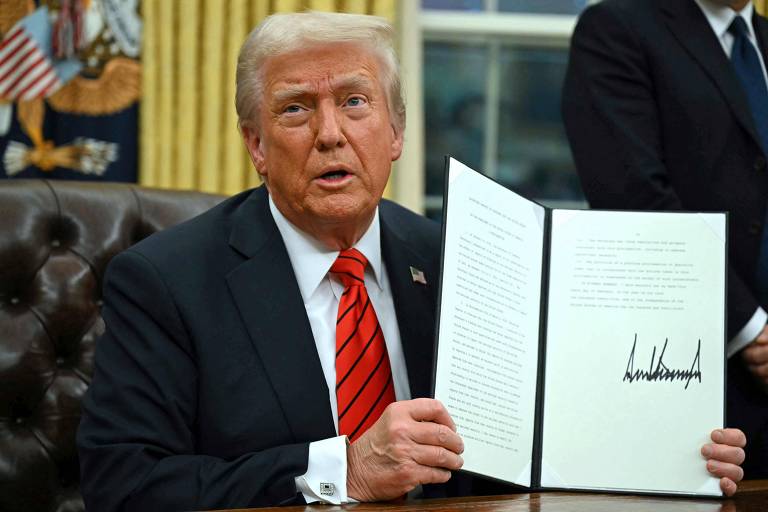 Um homem com cabelo loiro e uma gravata vermelha está sentado em uma mesa， segurando um documento em uma das mãos. Ele parece estar em um escritório， com uma bandeira americana ao fundo. O documento tem uma assinatura visível na parte inferior.
