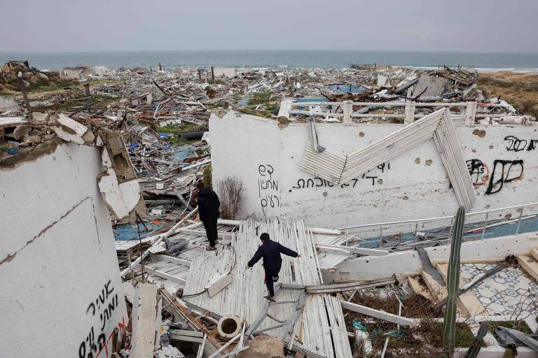 A imagem mostra uma área devastada por um desastre natural， com edifícios em ruínas e destroços espalhados. Duas pessoas estão caminhando sobre os escombros， com o mar visível ao fundo. O céu está nublado， sugerindo um clima sombrio