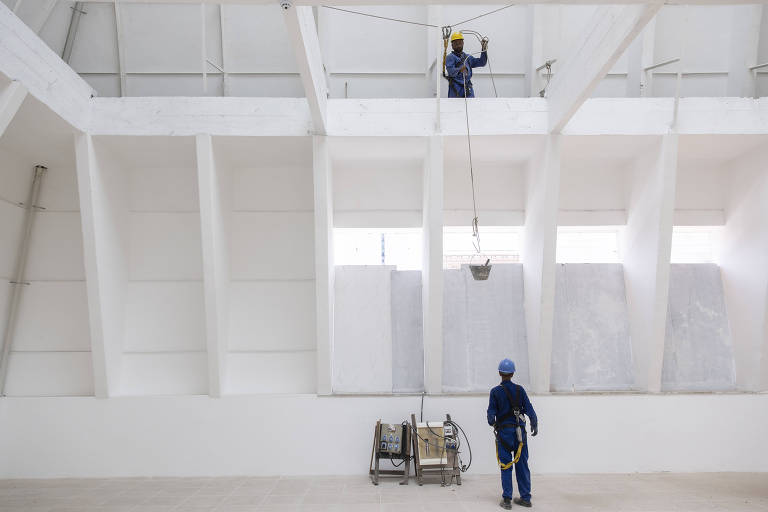 A imagem mostra dois trabalhadores em um ambiente industrial. Um deles está em uma plataforma elevada， utilizando um equipamento para levantar um balde， enquanto o outro está no chão， observando. O ambiente é amplo， com paredes brancas e uma estrutura de suporte visível.
