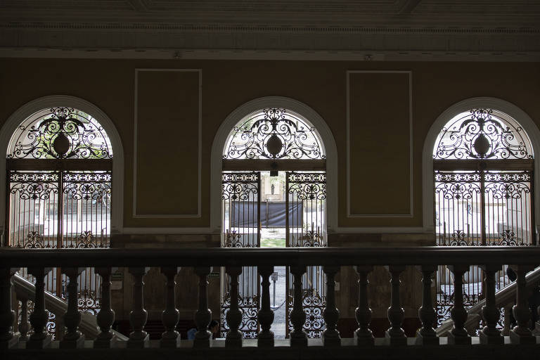 A imagem mostra três janelas arqueadas com grades decorativas em um ambiente interno. As janelas estão dispostas em uma parede clara， e a luz natural entra， iluminando o espaço. Abaixo das janelas， há um corrimão escuro que complementa a arquitetura do local.
