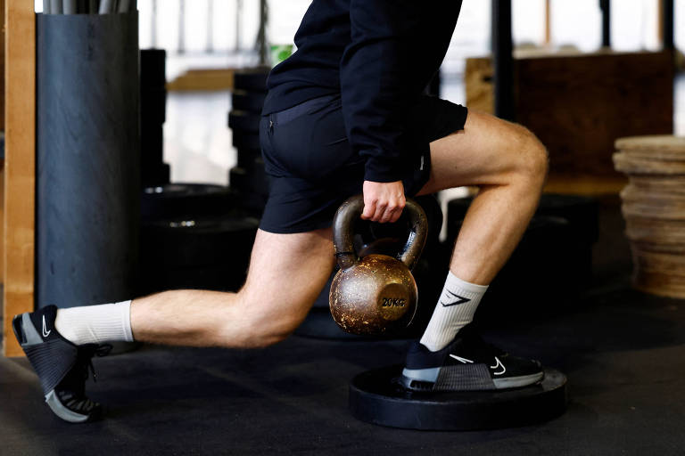 A imagem mostra uma pessoa realizando um exercício de agachamento com um kettlebell. A pessoa está em uma posição de avanço， segurando um kettlebell de 20 kg com a mão direita. Está vestindo uma camiseta de manga longa preta e shorts pretos， além de meias brancas e tênis escuros. O fundo é de uma academia， com pesos e equipamentos visíveis.