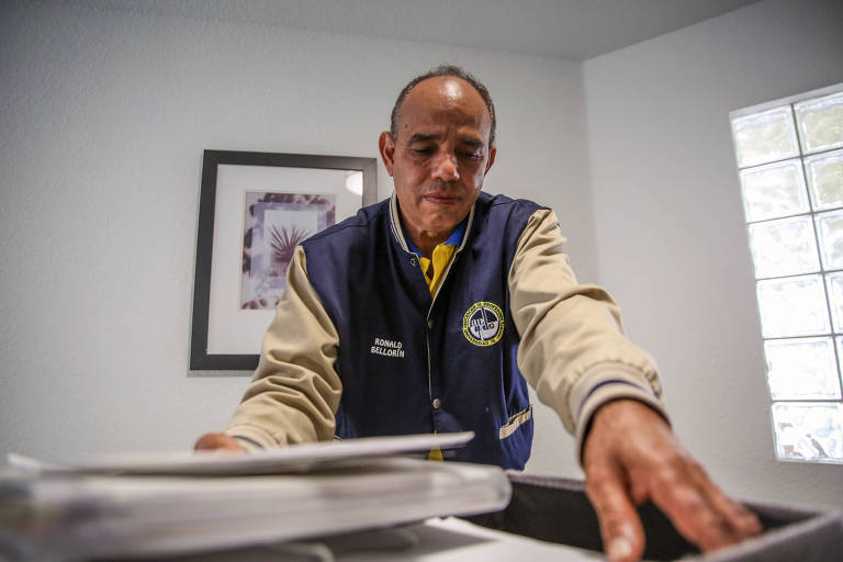 Um homem está em um ambiente interno， aparentemente organizando documentos sobre uma mesa. Ele usa uma jaqueta azul com detalhes em bege e uma camisa amarela por baixo. Ao fundo， há uma parede branca e uma janela com vidro. Uma obra de arte emoldurada está pendurada na parede ao lado.