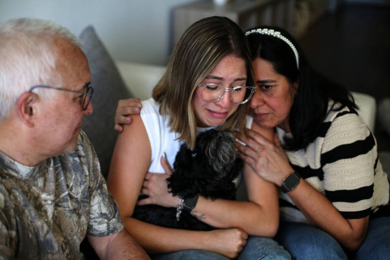 A imagem mostra três pessoas sentadas em um sofá. Uma jovem mulher， com cabelo liso e óculos， está chorando enquanto segura um cachorro preto em seus braços. Ela é confortada por duas pessoas ao seu lado: um homem idoso com cabelo grisalho e uma mulher com cabelo escuro， que está usando uma blusa listrada. A atmosfera é de apoio emocional.