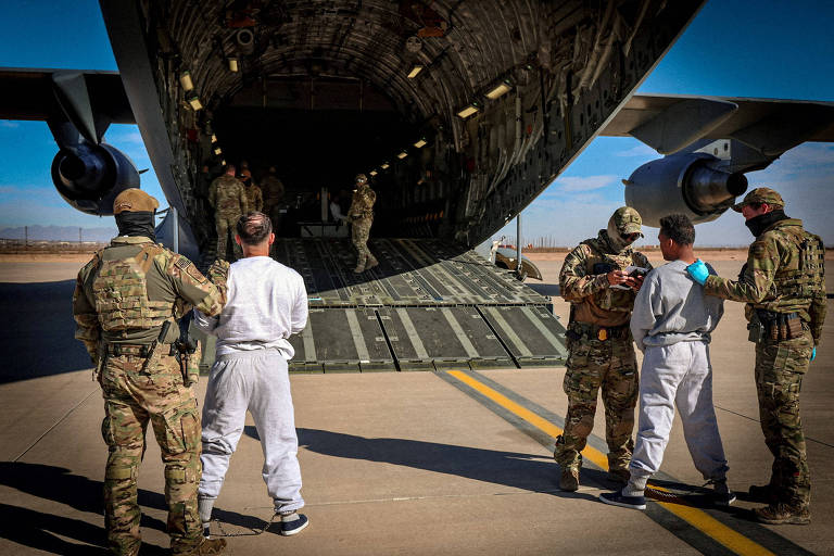 A imagem mostra um grupo de soldados em um aeroporto， próximos a um grande avião de transporte militar. Dois indivíduos， aparentemente em trajes de prisioneiro， estão sendo acompanhados por os soldados. O fundo apresenta uma pista de pouso e o céu é claro， com algumas nuvens.
