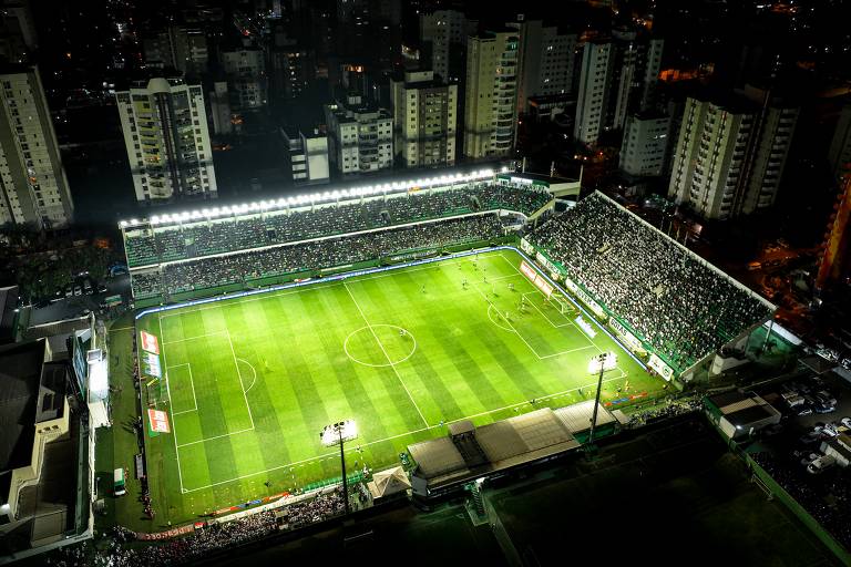 Imagem aérea de um estádio de futebol iluminado à noite， com um campo verde bem cuidado. O estádio está cheio de torcedores， e há várias luzes acesas ao redor. No campo， jogadores estão em ação， e há bandeiras e anúncios visíveis nas laterais. Ao fundo， há prédios altos e iluminados.