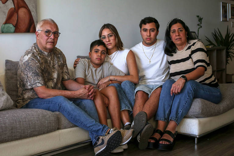 A imagem mostra uma família sentada em um sofá. À esquerda， um homem idoso com óculos e camisa camuflada. Ao seu lado， um menino de camiseta cinza. No centro， uma jovem mulher com óculos e camiseta branca， que está abraçando o menino. À direita， um jovem homem com camiseta branca e corrente no pescoço， e uma mulher com blusa listrada e cabelo longo. O ambiente é claro， com uma parede decorada e plantas ao fundo.