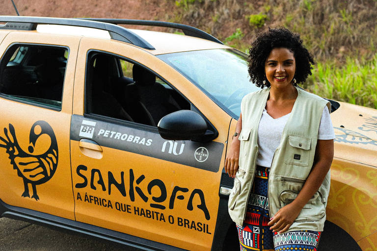 A imagem mostra uma mulher sorridente ao lado de um carro amarelo com detalhes gráficos. Ela usa um colete claro e uma camiseta branca， além de uma saia com estampas coloridas. O carro possui a inscrição 039;SANKOFA039; e a frase 039;A ÁFRICA QUE HABITA O BRASIL039;， junto com logotipos de empresas como Petrobras e Nu. O fundo é uma área verde com vegetação.