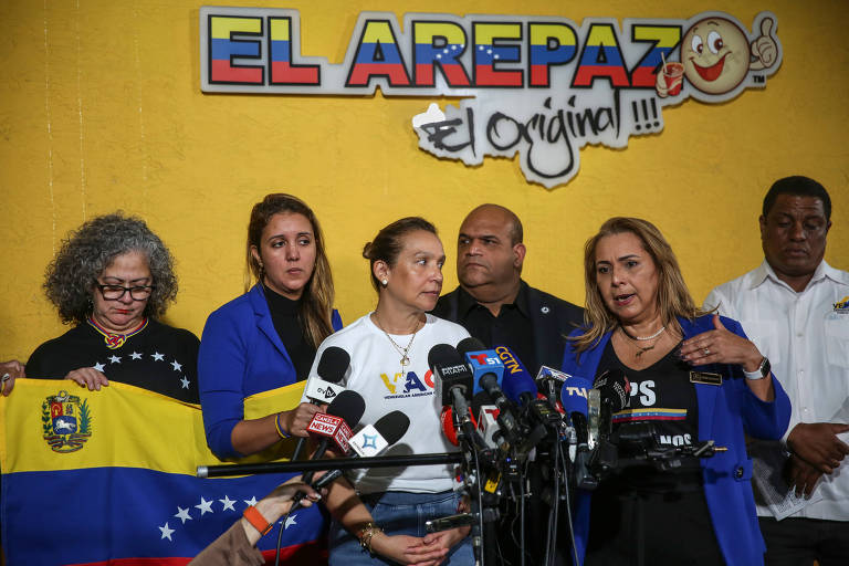 A imagem mostra um grupo de pessoas em uma conferência de imprensa. No centro， uma mulher fala ao microfone， enquanto outras mulheres e um homem a escutam. Uma delas segura uma bandeira da Venezuela. O fundo é amarelo com o nome 039;EL AREPAZO039; em destaque. Vários microfones estão posicionados à frente do grupo.