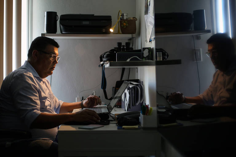 A imagem mostra um homem sentado em uma mesa de escritório， concentrado em seu laptop. Ele usa óculos e está vestido com uma camisa de manga longa. A iluminação é suave， com luz natural entrando pela janela à esquerda， criando um contraste entre a luz e a sombra. No fundo， há prateleiras com objetos como uma impressora， uma cesta e outros itens de escritório.