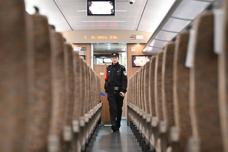 A imagem mostra o interior de um trem com assentos organizados em fileiras. Um funcionário， vestido com uniforme， caminha pelo corredor central entre os assentos. O ambiente é bem iluminado， e há telas visíveis ao fundo.