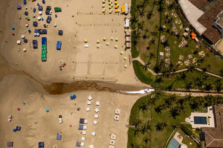 Imagem aérea de uma praia com várias barracas e guarda-sóis coloridos. O areal apresenta áreas com cadeiras de praia e uma faixa de água que parece ser um pequeno riacho. À direita， há uma área gramada com palmeiras e espreguiçadeiras， além de uma piscina visível. A imagem mostra uma grande quantidade de pessoas aproveitando o espaço.