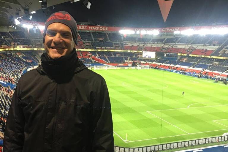 Um homem sorridente está posando em um estádio de futebol. Ele usa um casaco escuro e um gorro. Ao fundo， vê-se o campo de futebol iluminado， com arquibancadas e uma atmosfera de jogo. A iluminação do estádio é intensa， e há bandeiras penduradas no teto