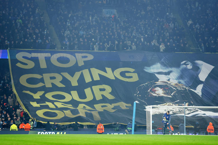 A imagem mostra uma grande bandeira de torcida em um estádio de futebol. A bandeira possui a frase em letras grandes e amarelas: 039;STOP CRYING YOUR HEART OUT039;. Ao fundo， é possível ver a arquibancada cheia de torcedores e um goleiro em frente ao gol.
