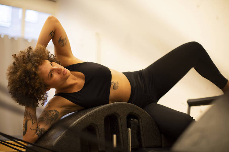 A imagem mostra uma pessoa praticando Pilates em um equipamento conhecido como reformer. A pessoa， que tem cabelos encaracolados e usa uma roupa de treino preta， realiza um exercício que envolve torção e alongamento do tronco， destacando a flexibilidade e o controle muscular. O cenário é uma sala clara， possivelmente um estúdio de Pilates， com paredes brancas que contribuem para um ambiente calmo e focado. A expressão da pessoa sugere concentração intensa enquanto ela executa o movimento， ilustrando a natureza desafiadora e envolvente do Pilates.







