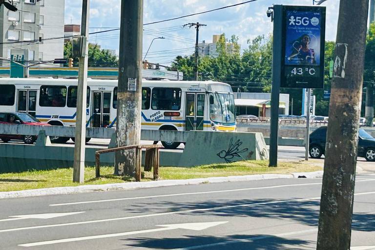 A imagem mostra um ônibus urbano estacionado em uma parada， com um painel publicitário ao fundo. O cenário é urbano， com árvores e postes visíveis. O céu está claro e ensolarado.