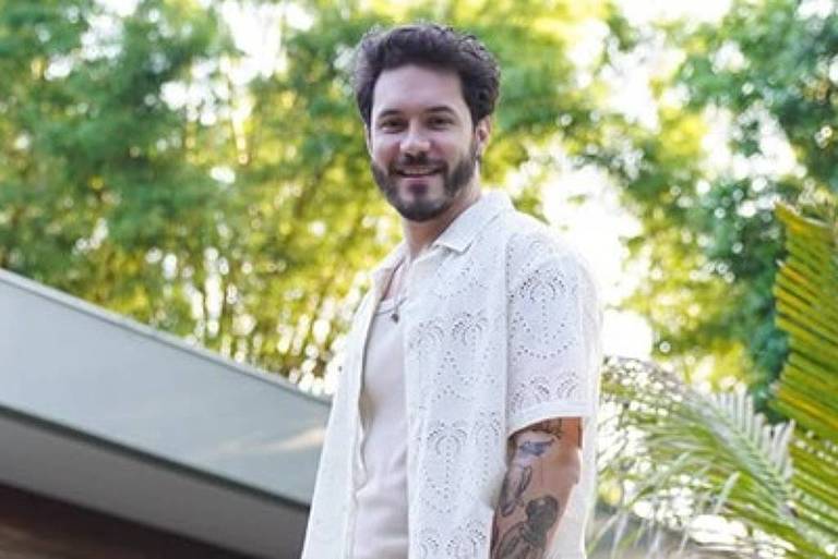 Em foto colorida, homem todo de branco posa para foto em frente a uma casa