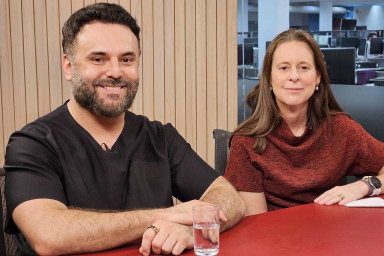 Duas pessoas sentadas em uma mesa. À esquerda， um homem com barba e cabelo escuro， vestindo uma camiseta preta. À direita， uma mulher com cabelo castanho claro， usando um suéter marrom. Ambos estão sorrindo e parecem estar em um ambiente de trabalho ou estúdio.