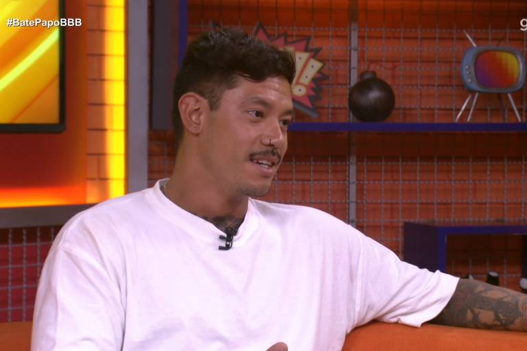 Um homem com cabelo curto e um bigode está sentado em um sofá laranja, usando uma camiseta branca. Ele parece estar conversando e gesticulando. Ao fundo, há uma parede laranja com prateleiras que exibem uma bola decorativa e uma televisão antiga. O ambiente é iluminado e moderno.
