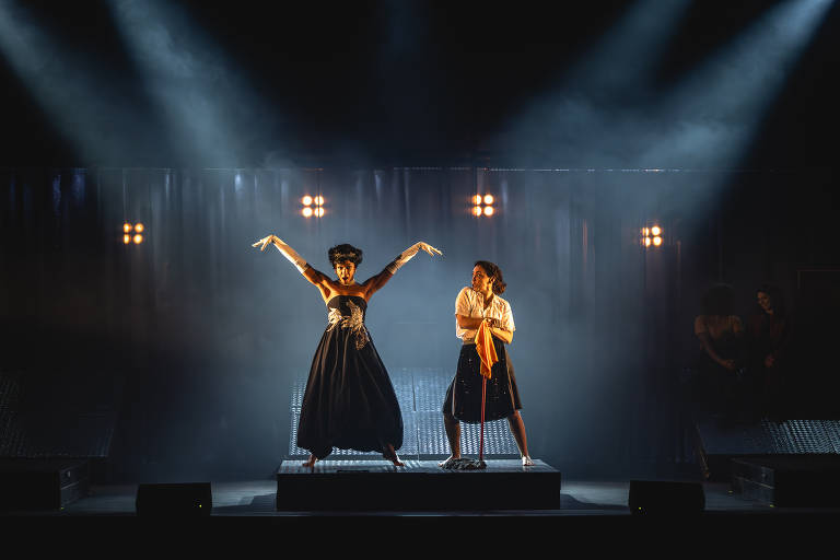 A imagem mostra duas performers em um palco iluminado. À esquerda， uma mulher está vestida com um vestido longo preto e luvas brancas， com os braços levantados em uma pose expressiva. À direita， outra mulher está vestida com uma camisa branca e uma saia preta， segurando um pano laranja e olhando para a primeira mulher. O fundo é escuro， com luzes brilhantes e uma leve névoa， criando uma atmosfera dramática.