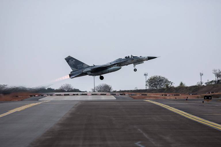 Um avião de caça está decolando de uma pista， com chamas saindo dos motores. O céu está nublado e a pista é de concreto， com marcas amarelas. Ao fundo， há árvores e estruturas de apoio.