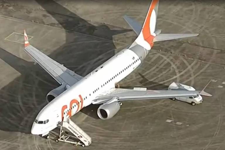 Imagem de um avião da companhia aérea Gol， modelo Boeing 737， estacionado em uma pista de aeroporto. O avião possui uma pintura branca com detalhes em laranja. Há uma escada de embarque conectada à porta dianteira do avião e um veículo de serviço próximo à fuselagem.