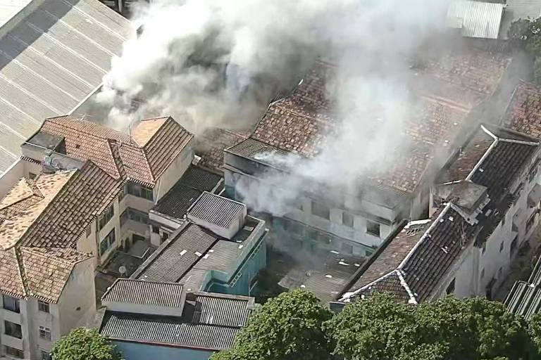 A imagem mostra uma área urbana com várias construções， onde há fumaça saindo de um dos edifícios. Os telhados são de cerâmica e alguns estão danificados. A fumaça é densa e se espalha pelo ar， indicando um incêndio em andamento. Árvores estão visíveis na parte inferior da imagem.