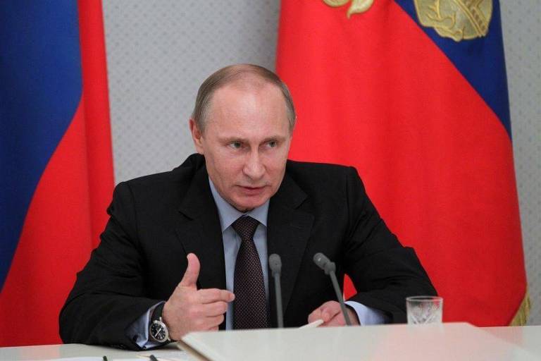 Um homem está sentado à mesa， gesticulando com a mão direita enquanto fala. Ele usa um terno escuro e uma camisa clara. Ao fundo， há duas bandeiras da Rússia， uma com as cores azul， branco e vermelho， e a outra com o brasão da Rússia. Há um copo de vidro sobre a mesa.