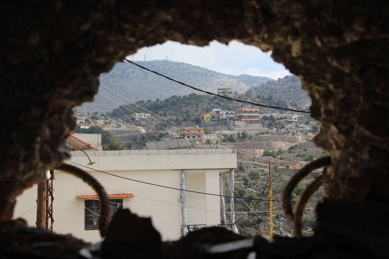 A imagem mostra uma vista através de uma abertura em uma parede de pedra. Ao fundo， há uma paisagem montanhosa com várias casas construídas em uma encosta. A parte inferior da imagem apresenta fios elétricos e uma estrutura de concreto. O céu está nublado， e a vegetação é visível nas montanhas.