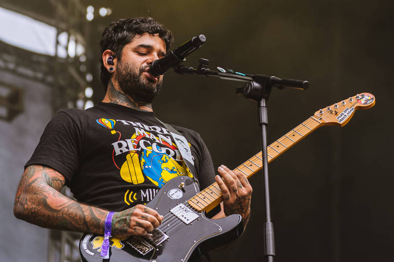 Um músico está se apresentando em um palco, segurando uma guitarra elétrica preta. Ele usa uma camiseta preta com uma estampa colorida. O músico tem tatuagens visíveis nos braços e está cantando em um microfone. O fundo é iluminado com luzes amarelas e há uma leve névoa no ar.