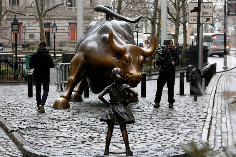 A imagem mostra a famosa escultura do Touro de Wall Street， uma grande escultura de bronze de um touro， em um espaço público. Em frente ao touro， está a escultura da Menina Corajosa， uma figura de bronze de uma menina com as mãos na cintura， encarando o touro. Há pessoas ao redor， incluindo um homem com um casaco escuro e um fotógrafo. O ambiente é urbano， com calçadas de pedras e árvores ao fundo.
