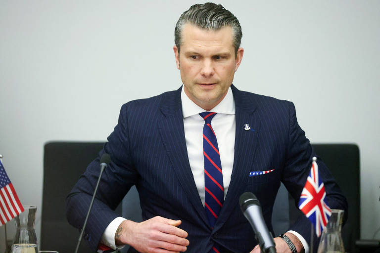 Um homem em um terno listrado azul escuro com uma gravata vermelha e branca está sentado à mesa. Ele parece estar em um ambiente formal， com bandeiras dos Estados Unidos e do Reino Unido visíveis na mesa. O homem tem cabelo grisalho e está olhando para baixo， com as mãos sobre a mesa.