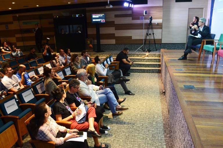 A imagem mostra um auditório com várias pessoas sentadas assistindo a uma apresentação. No palco， há um palestrante e uma pessoa ao lado dele. O público está atento， com algumas pessoas segurando papéis ou dispositivos. O ambiente é bem iluminado e possui um design moderno， com painéis de madeira nas paredes.
