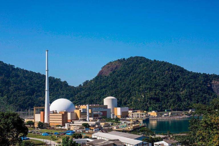 A imagem mostra uma usina nuclear localizada em uma área montanhosa， com um grande reator de forma esférica e uma chaminé alta. Ao fundo， há montanhas cobertas de vegetação densa e um corpo d039;água à direita. A usina é composta por várias estruturas de diferentes tamanhos e cores， com um céu azul claro ao fundo.