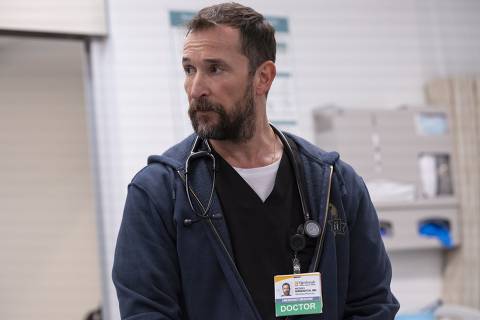 Robby and team work to pinpoint Nicks condition. (Warrick Page/MAX) *** Local Caption *** Um homem com barba e cabelo curto, vestindo um jaleco escuro e um moletom azul, está em um ambiente hospitalar. Ele usa um estetoscópio ao redor do pescoço e tem um crachá visível que indica que ele é um médico de emergência. O fundo mostra equipamentos hospitalares e uma cortina.