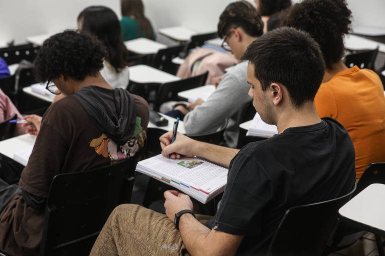 A imagem mostra um grupo de estudantes sentados em uma sala de aula. Eles estão concentrados em suas atividades， com alguns escrevendo em cadernos. A maioria dos alunos está de costas， e é possível ver diferentes estilos de roupas. O ambiente é simples， com cadeiras e mesas dispostas em fileiras.