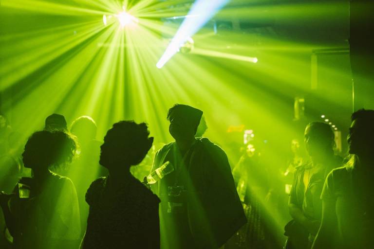 A imagem mostra um ambiente de festa com várias pessoas dançando. A iluminação é predominantemente verde, com feixes de luz que se espalham pelo espaço. As silhuetas das pessoas são visíveis, mas os detalhes faciais não são discerníveis devido à iluminação. O ambiente parece animado e festivo.