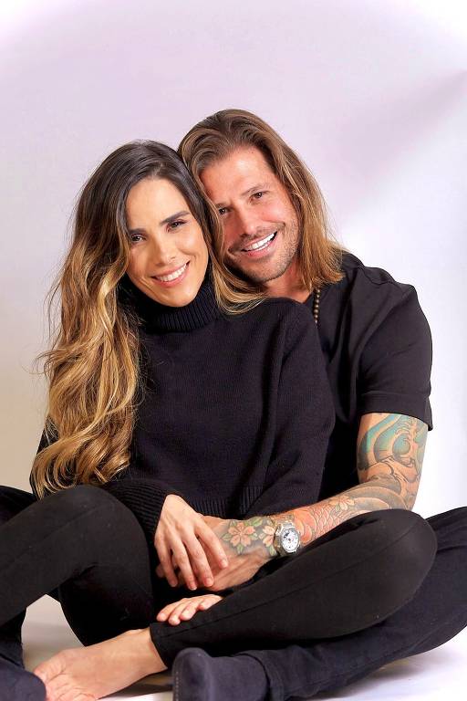 A imagem mostra um casal sentado no chão, com as pernas cruzadas. A mulher está à frente, usando um suéter preto e sorrindo, enquanto o homem está atrás dela, com os braços envolvendo-a. Ele usa uma camiseta preta e tem tatuagens visíveis nos braços. Ambos têm cabelos longos e estão descalços, com um fundo claro.