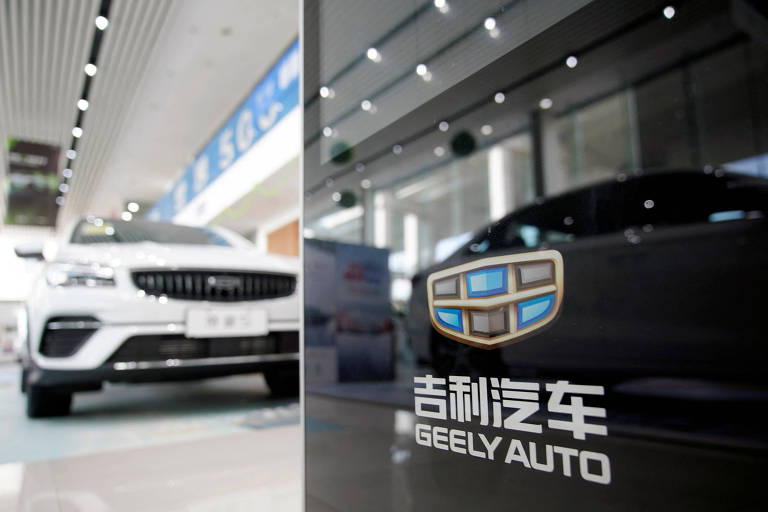 A imagem mostra um showroom da Geely Auto， com um carro branco em destaque à esquerda e um carro preto à direita. O logotipo da Geely Auto está visível na parte inferior direita da imagem， com caracteres em chinês e a tradução 039;Geely Auto039; em inglês. O ambiente é moderno， com iluminação clara e paredes em tons neutros.
