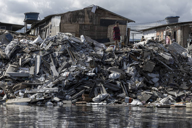 A imagem mostra uma grande pilha de lixo e entulho em uma área urbana， com uma construção ao fundo. Duas pessoas estão em cima da pilha de resíduos， que inclui materiais como metal e plástico. O céu está nublado， e a cena reflete uma situação de poluição e descaso ambiental.